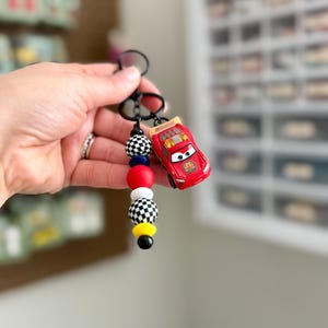 May include: A red toy car keychain with a black keyring and a beaded accent. The beads are in red, white, black, and yellow. The toy car is a Lightning McQueen character.