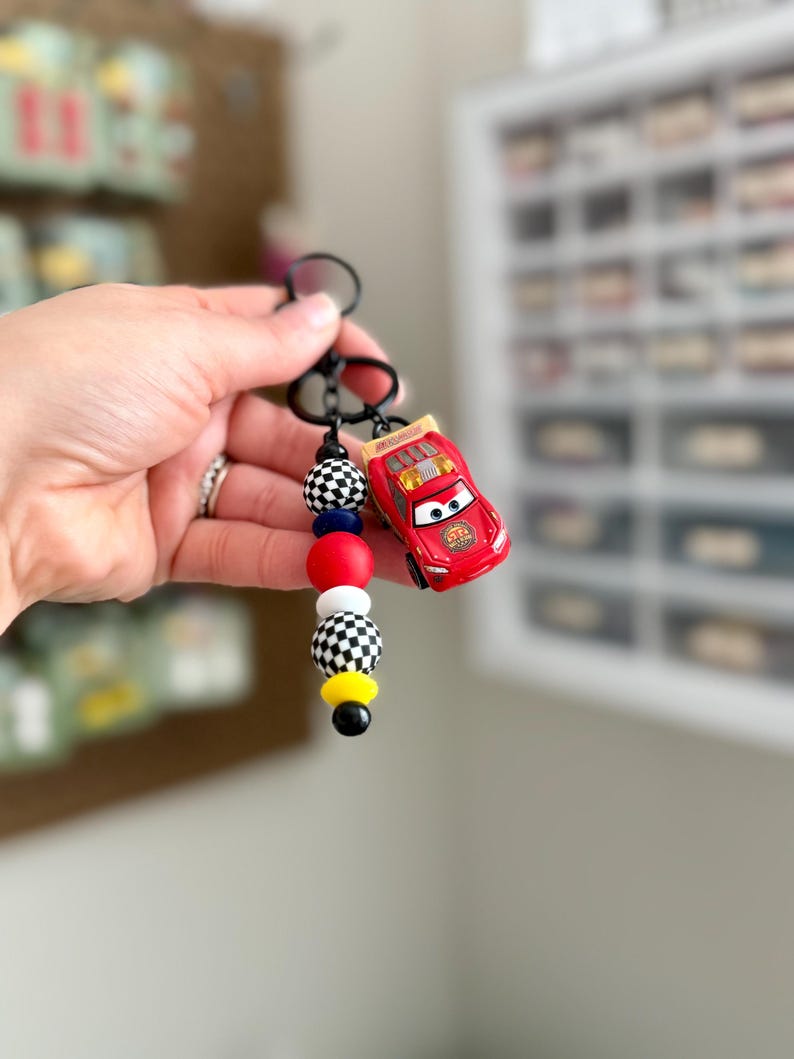 May include: A red toy car keychain with a black keyring and a beaded accent. The beads are in red, white, black, and yellow. The toy car is a Lightning McQueen character.