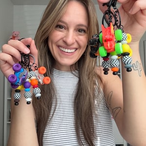 May include: Two keychains featuring miniature monster truck toys attached to black metal rings. The keychains are decorated with colorful silicone beads in solid orange, purple, yellow, and black-and-white checkered patterns, suspended from black clips.