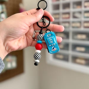 May include: A keychain featuring a small, light blue race car toy with the number 43. The keychain also includes a series of decorative beads in red, white, black, and checkered patterns, attached to a black metal ring.