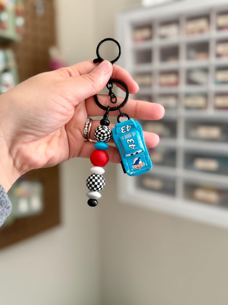 May include: A keychain featuring a small, light blue race car toy with the number 43. The keychain also includes a series of decorative beads in red, white, black, and checkered patterns, attached to a black metal ring.