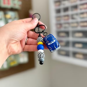 May include: A blue toy car keychain with a black keyring and a beaded accent. The beads are black and white checkered, yellow, blue, gray, and black. The toy car is a miniature version of a race car.