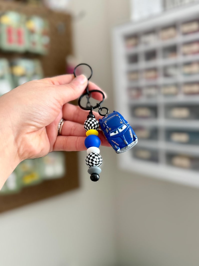 May include: A blue toy car keychain with a black keyring and a beaded accent. The beads are black and white checkered, yellow, blue, gray, and black. The toy car is a miniature version of a race car.