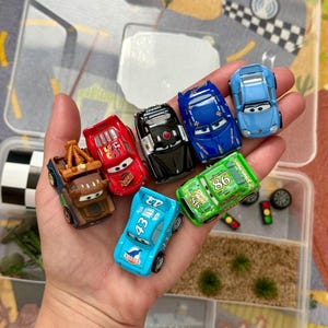 May include: A collection of toy cars in various colors, including red, blue, and green, held in a clear plastic container. The cars feature character designs and racing numbers. The container has multiple compartments with decorative elements.
