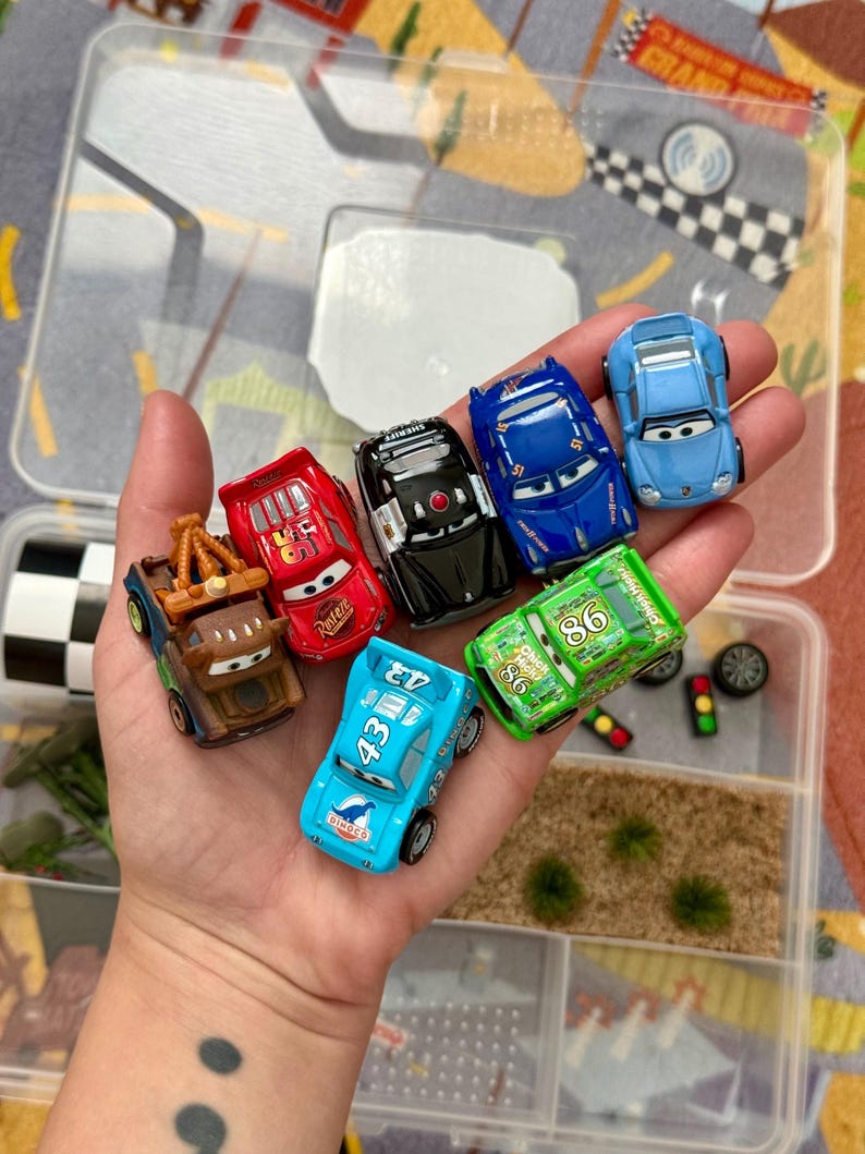 May include: A collection of toy cars in various colors, including red, blue, and green, held in a clear plastic container. The cars feature character designs and racing numbers. The container has multiple compartments with decorative elements.