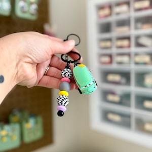May include: A keychain featuring a green toy car and a beaded charm. The charm includes black and white checkered beads, gray, yellow, pink, and black beads. The keychain is held in a person's hand.