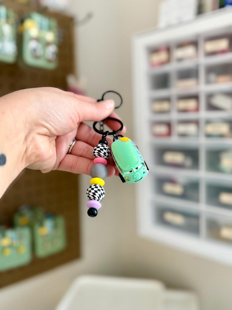 May include: A keychain featuring a green toy car and a beaded charm. The charm includes black and white checkered beads, gray, yellow, pink, and black beads. The keychain is held in a person's hand.
