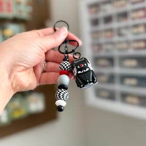 May include: A black keychain with a small black car charm and a beaded accent. The beads include red, gray, black, and white, with checkered and solid patterns. The car charm has red details.