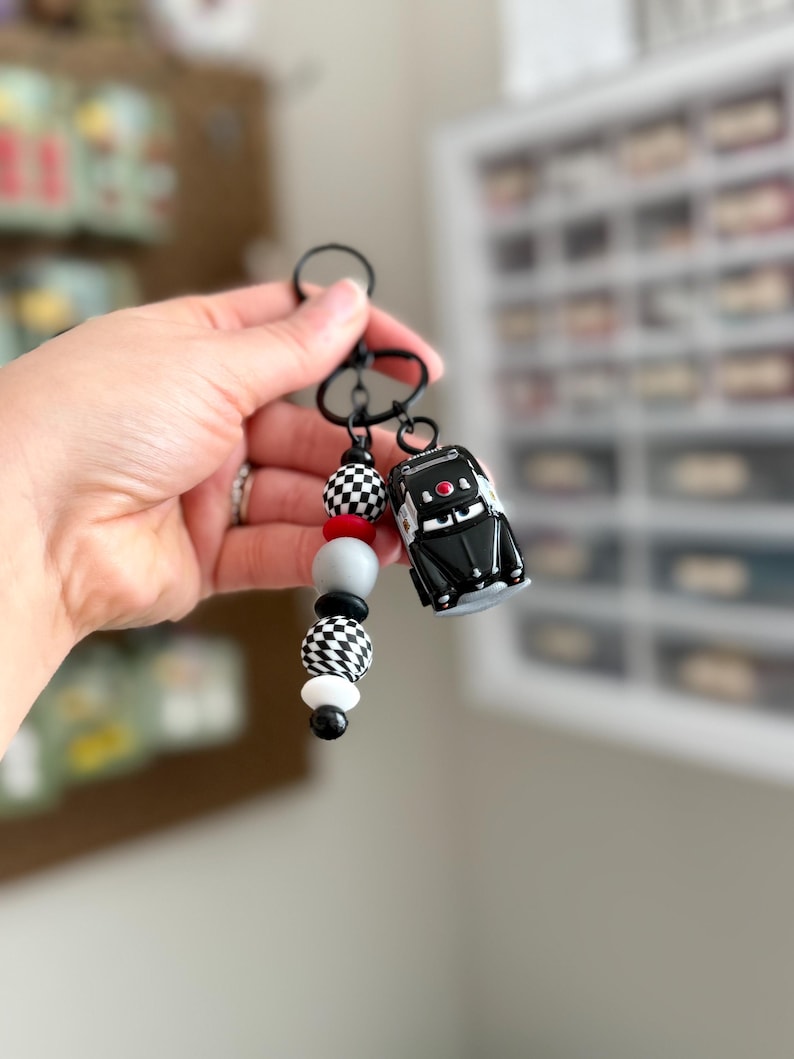 May include: A black keychain with a small black car charm and a beaded accent. The beads include red, gray, black, and white, with checkered and solid patterns. The car charm has red details.