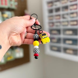 May include: A black keychain with a yellow toy car charm and a series of colorful beads. The beads include black and white checkered, yellow, red, and black. The keychain is held up by a hand.
