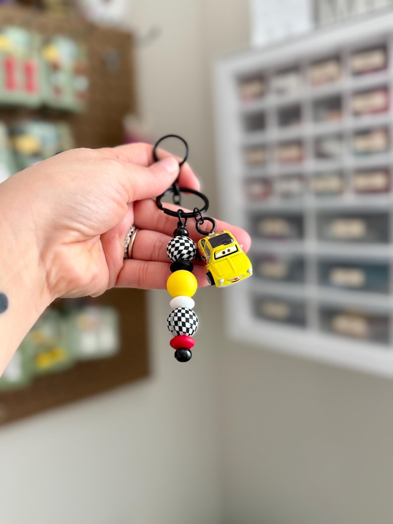 May include: A black keychain with a yellow toy car charm and a series of colorful beads. The beads include black and white checkered, yellow, red, and black. The keychain is held up by a hand.