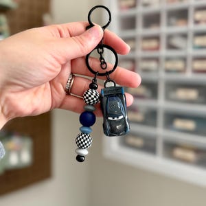 May include: A black keychain with a miniature blue race car and a beaded charm. The charm features black and white checkered beads, blue and gray beads, and a black bead. The keychain is held by a hand.