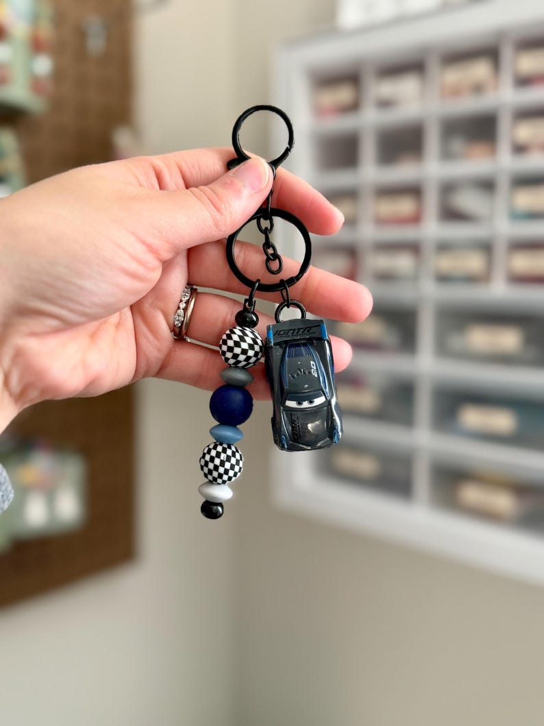 May include: A black keychain with a miniature blue race car and a beaded charm. The charm features black and white checkered beads, blue and gray beads, and a black bead. The keychain is held by a hand.
