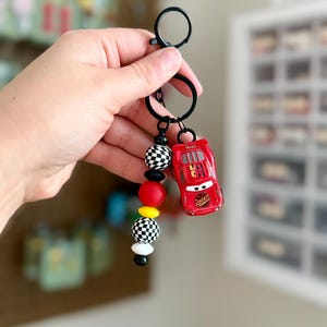 May include: A red toy car keychain with the number 95 on the side, attached to a black keyring. The keychain also features a strand of black and white checkered beads, red, yellow, and white beads.