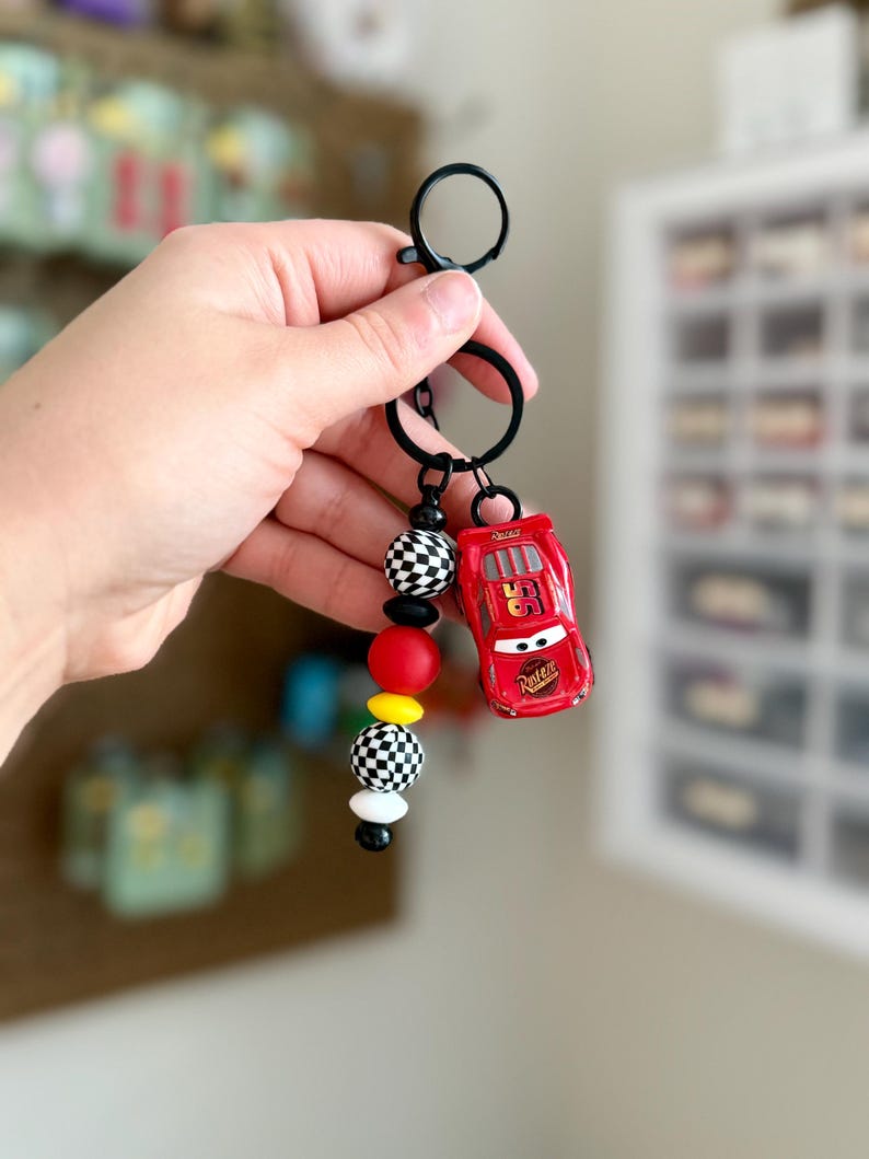 May include: A red toy car keychain with the number 95 on the side, attached to a black keyring. The keychain also features a strand of black and white checkered beads, red, yellow, and white beads.