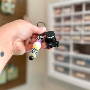 May include: A black car-shaped keychain with a beaded strand. The beads are white, black, yellow, and lavender. The car has a smiling face. The keychain is held up against a blurred background.