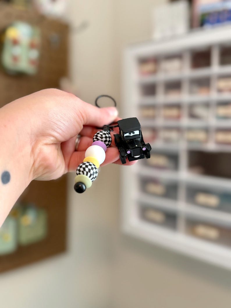 May include: A black car-shaped keychain with a beaded strand. The beads are white, black, yellow, and lavender. The car has a smiling face. The keychain is held up against a blurred background.