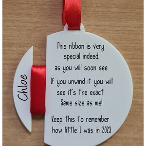 May include: White round ornament with a red ribbon attached. The ornament has the text "Chloe" on one side and the text "This ribbon is very special indeed, as you will soon see. If you unwind it you will see it's the exact Same size as me! Keep this to remember how little I was in 2023" on the other side.