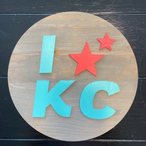 May include: A round wooden sign with the words "I ❤️ KC" in teal and red. The letters and heart are raised from the wooden surface. The background is a light brown color.