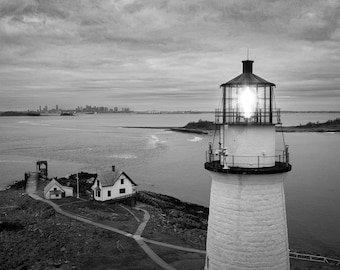May include: A black and white photo of a lighthouse with a bright light shining from the top. The lighthouse is on a small island with a small house and a path leading to it. The city skyline is visible in the distance.