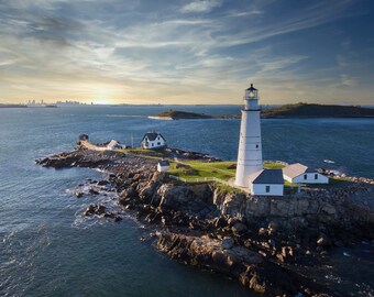 May include: A white lighthouse stands tall on a rocky island, with a smaller white house nearby. The ocean stretches out in the background, with a city skyline visible in the distance.
