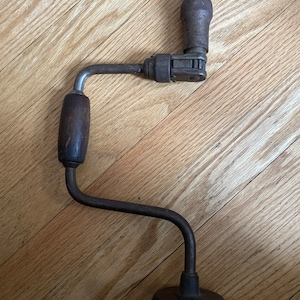 May include: A vintage hand-crank drill with a wooden handle and a metal crank. The drill is made of metal and has a black finish. It is a useful tool for drilling holes in wood.