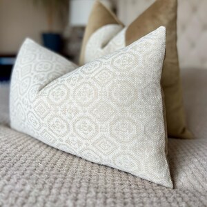 May include: Two decorative pillows on a bed. The front pillow has a cream-colored geometric pattern. The back pillow is a solid gold color. The pillows are resting on a beige bedspread. Home decor.