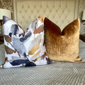 Può includere: Due cuscini decorativi. Uno presenta uno sfondo bianco con disegni astratti ad acquerello nei toni del marrone, grigio, oro e blu navy. L'altro cuscino è di colore marrone dorato pieno e morbido.
