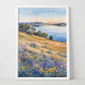 May include: Watercolor painting of a serene landscape featuring a lake and rolling hills. The foreground is filled with wildflowers in shades of yellow, purple, and lavender. The sky transitions from blue to a soft yellow, suggesting sunrise or sunset. The artwork is framed in a white border.