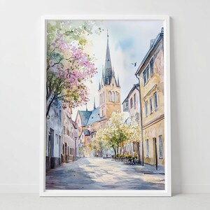 Puede incluir: Pintura a la acuarela de una escena de calle europea con un alto campanario de iglesia. Los edificios son en tonos beige y marrón, con árboles en flor. La obra de arte está enmarcada en un marco blanco.
