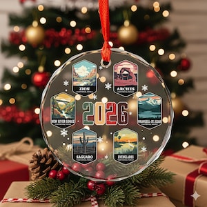 May include: A clear, round Christmas ornament with a red ribbon. The ornament features illustrations of national parks, including Zion, Arches, and Everglades, with the year 2026 in the center. Snowflakes and text are also included.