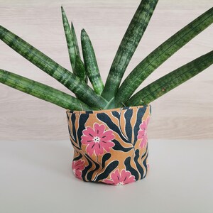 Peut inclure: Une plante serpent en pot avec des feuilles vertes dans un pot en tissu à motif floral. Le pot a un fond brun avec des fleurs roses et des feuilles noires.