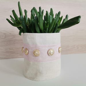 Peut inclure: Une plante succulente dans un pot en tissu blanc avec une bande rose et des boutons dorés.