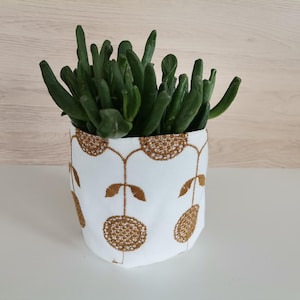 Peut inclure: Un pot en tissu blanc avec un motif floral doré, contenant une plante succulente aux feuilles vertes.