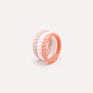 Può includere: Un set di tre anelli impilati caratterizzati da una texture a corda intrecciata. Gli anelli sono colorati in rosa chiaro, bianco e un vivace arancione corallo. Ogni anello ha una forma circolare uniforme e un motivo intrecciato costante.