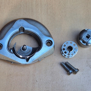 May include: A silver-toned metal sewing machine rotary hook assembly with the text SIMANCO 2515, a circular metal bobbin, a metal bobbin case with a small lever, and two threaded metal screws arranged on a flat surface.