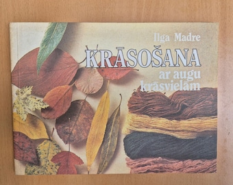 May include: A book cover featuring the title "KRĀSOŠANA" with text in Latvian, and the author's name, Ilga Madre. The cover is decorated with colorful autumn leaves and a stack of yarn in various shades of brown, yellow, and blue.