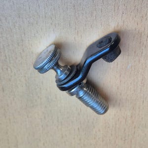 Puede incluir: Un componente de hardware metálico que presenta un perno roscado, un soporte angular negro con una abertura circular y una perilla de ajuste moleteada de tono plateado. El conjunto incluye una pequeña arandela de goma negra entre el perno y el soporte.