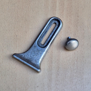 May include: A silver-colored metal part with a slotted design and a small, round knob. The larger piece has a flared base and a rectangular opening. The knob has a ridged edge. Both pieces are on a light brown wooden surface.