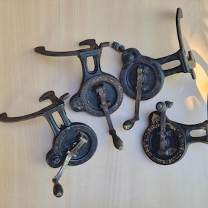 K&ouml;nnte beinhalten: Vier schwarze N&auml;hmaschinen-Handkurbeln aus Gusseisen mit goldfarbenen Blumenverzierungen auf den runden Radfl&auml;chen. Jeder Mechanismus verf&uuml;gt &uuml;ber einen geschwungenen Montagearm und einen kleinen Holzgriff am drehbaren Metallkurbelarm.