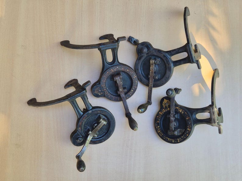 K&ouml;nnte beinhalten: Vier schwarze N&auml;hmaschinen-Handkurbeln aus Gusseisen mit goldfarbenen Blumenverzierungen auf den runden Radfl&auml;chen. Jeder Mechanismus verf&uuml;gt &uuml;ber einen geschwungenen Montagearm und einen kleinen Holzgriff am drehbaren Metallkurbelarm.