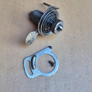 Könnte beinhalten: Eine silberfarbene mechanische Metallbaugruppe mit einem Gewindezylinder, einer Spiralfeder und einem flachen Hebel. Daneben liegt eine flache, kreisförmige Metallplatte mit einem kleinen Schlitz, einer Erhebung und dem Text C6245 sowie eine Schraube.