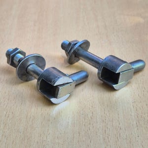Könnte beinhalten: Zwei silberfarbene Metallbeschläge, jeweils bestehend aus einem Gewindebolzen, einer flachen runden Unterlegscheibe und einem zylindrischen Kopf mit einem hervorstehenden Stift und einer quadratischen Aussparung. Die Metalloberflächen sind gebürstet.