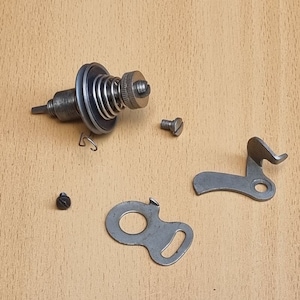 May include: A set of sewing machine parts, including a bobbin winder, a metal hook, and two screws. The parts are silver in color and are arranged on a light brown wooden surface.