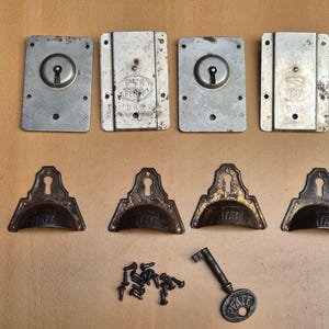 Könnte beinhalten: Ein Set aus Nähmaschinen-Ersatzteilen, bestehend aus vier rechteckigen Metallplatten mit Schlüssellöchern, vier dunklen Metall-Schubladengriffen mit PFAFF-Prägung, einem einzelnen Metallschlüssel mit PFAFF-Logo sowie einer kleinen Menge Schrauben.