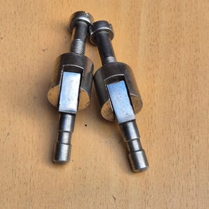 Könnte beinhalten: Zwei identische silberfarbene mechanische Metallbauteile, jedes mit einem zylindrischen Kopf mit geschlitzter Oberseite, einem zentralen Zylinderabschnitt mit einem rechteckigen hervorstehenden Arm und einem gestuften, schmalen zylindrischen Schaft.