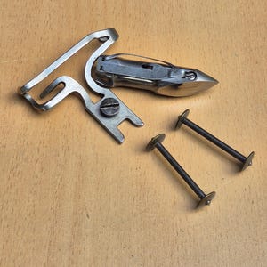 May include: A silver-toned metal sewing machine attachment featuring a curved guide arm and a central screw, accompanied by a separate metal shuttle component and two thin, dark metal bobbin-style pins with flat circular ends on each side.