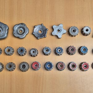 Pode incluir: Uma coleção de botões e puxadores de metal vintage em várias formas e tamanhos. Alguns botões têm detalhes em vermelho ou azul. Os itens estão dispostos em uma superfície marrom clara.