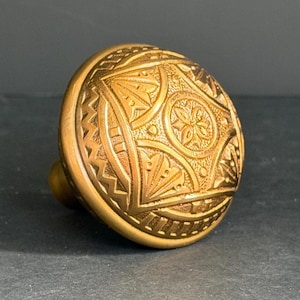 May include: A round, metallic gold-toned cabinet knob featuring an intricate, embossed geometric pattern on its face. The design includes a central circular motif surrounded by triangular segments and a textured border along the outer edge.