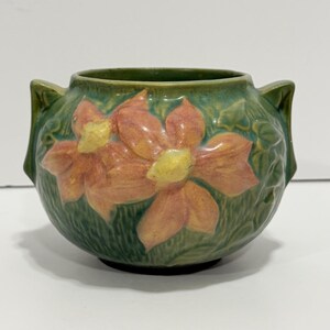May include: A round ceramic bowl with two small handles, featuring a green glazed finish and a raised relief design of two large orange flowers with yellow centers. The surface has a textured, leafy pattern surrounding the floral motif.